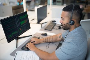 Man in a headset analyzing stock data on a computer monitor at his workspace.
